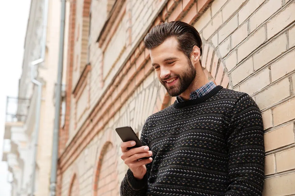 Young man smiling at phone screen with attractive mature woman's profile on a MILF dating app, evoking easy online connections for beginners.